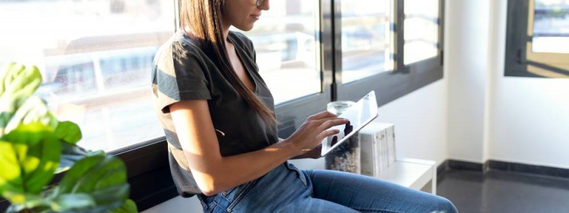 Jeune femme assise sur l'appui de fenêtre faisant des recherches sur sa tablette