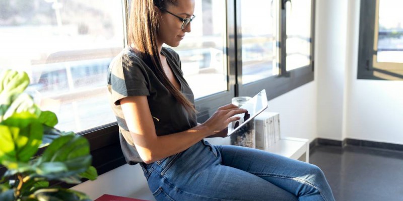 Jeune femme assise sur l'appui de fenêtre faisant des recherches sur sa tablette