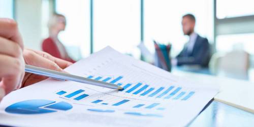 Gros plan sur la main d'un homme étudiant des graphiques avec deux personnes discutant en arrière-plan.